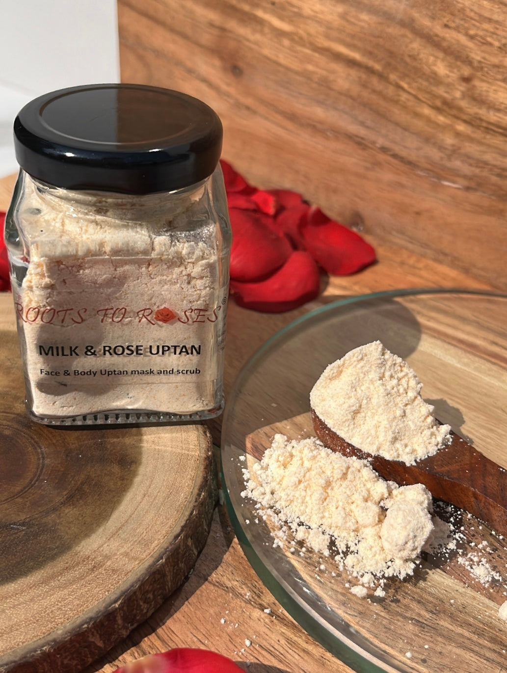 A glass jar labeled 'Milk Rose Uptan' with rose petals scattered around, and the product is shown spilling out of the jar onto a wooden surface.